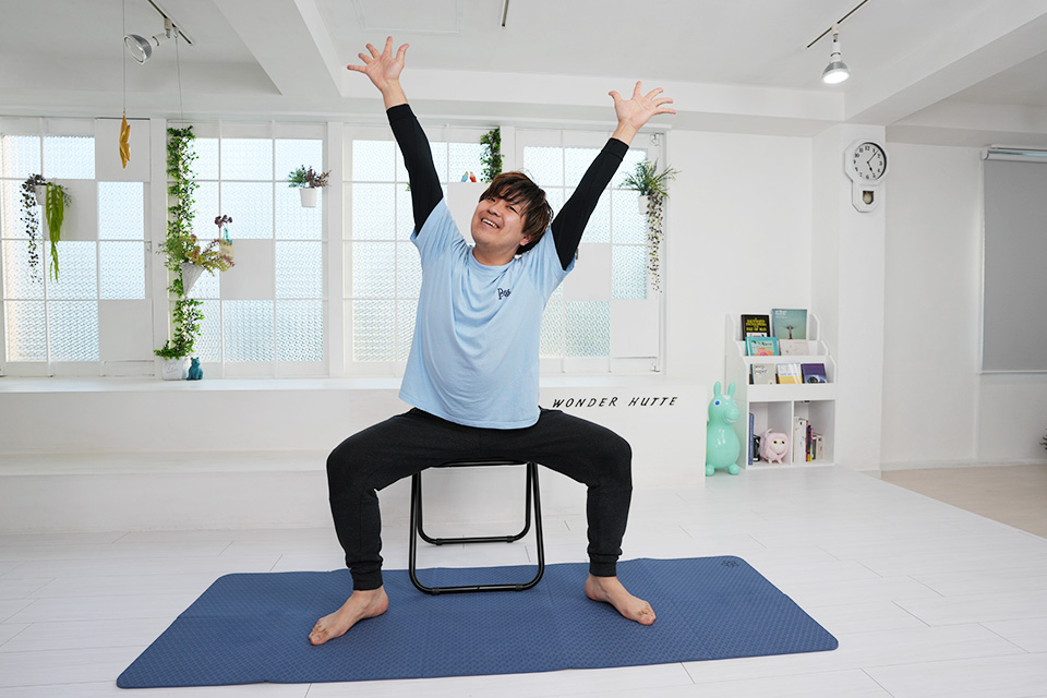 椅子に座ったまま、両腕を上に伸ばして笑っている参加者
